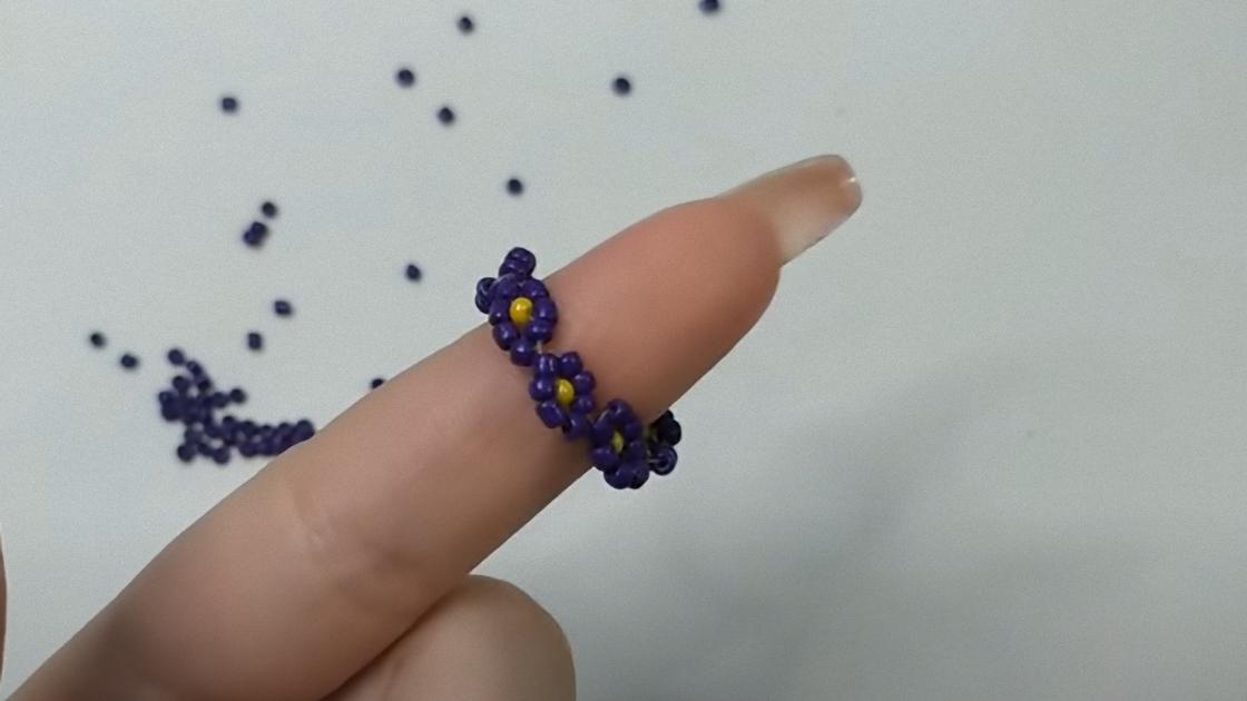 На пальце надето кольцо из фиолетового бисера в виде ромашек